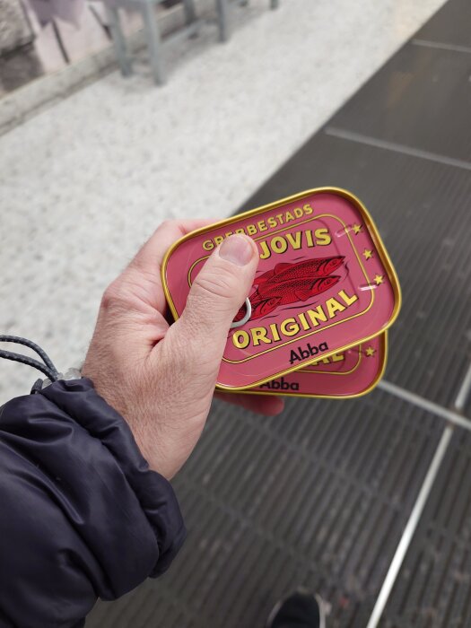 En hand som håller två små burkar ansjovisfiléer av märket Grebbestads, lämpliga för maträtten Janssons frestelse.