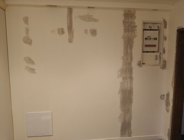 En vit vägg med spår av spackel, en inspektionslucka och elcentral synlig, förberedd för tapetsering.