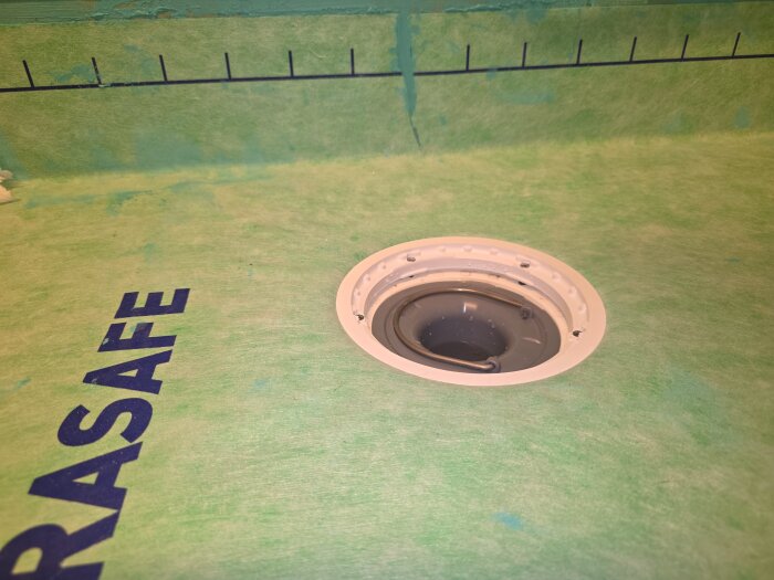 Avloppsbrunn på grön våtrumsmatta med texten "KERASAFE", luftbubblor och blått kladd syns nära väggen. Brunnen är omgiven av en tätning.