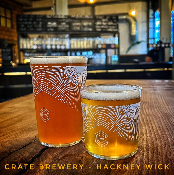Två ölglas på ett bord i Crate Brewery, Hackney Wick, med bakgrund av bryggeriets interiör och meny på väggen.