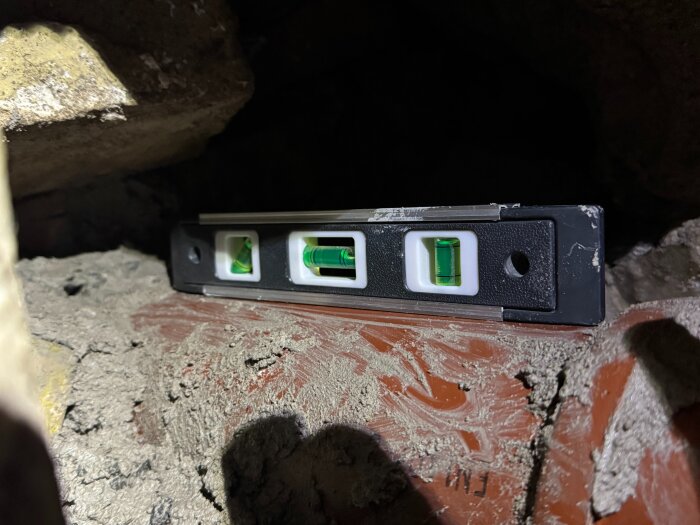 Vattenpass på ett lutande rör omgivet av stenar och fogskum i en källarvägg. Röret verkar luta bort från huset.