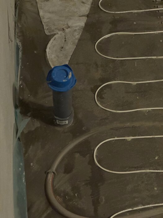 Badrumsgolv under renovering med el- och vattenburna golvvärmeslingor, flytspackel och synliga vattenrör nära golvbrunn.