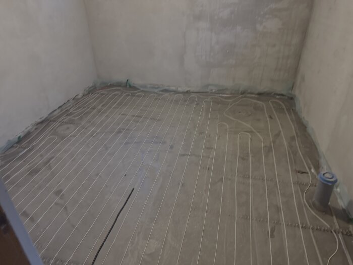 Badrumsgolvet i källaren med nyinstallerad golvvärme och flytspackel, vattenrör och brister i elinstallation nära avlopp synliga.