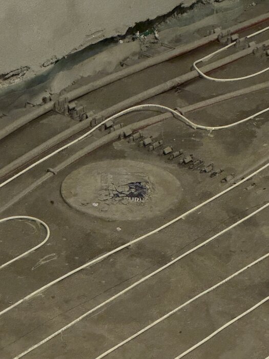 Golvvärmeinstallation med vatten- och elledningar nära golvbrunn i ett källarbadrum.