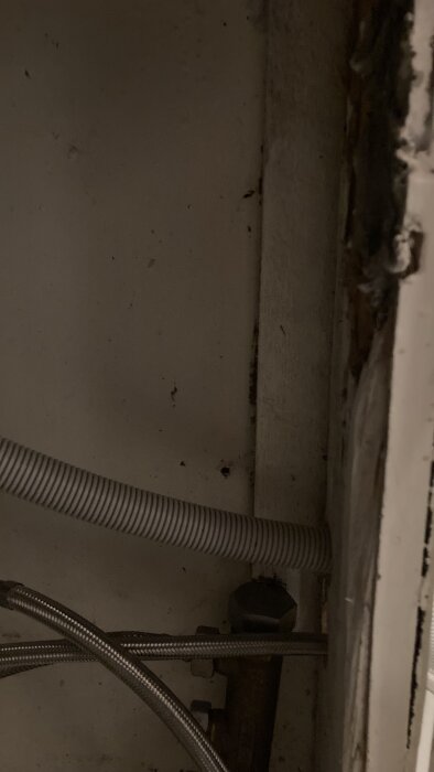 Mörk bild av ett trångt utrymme under en bänk med en vit ribbad slang och metallslangar.