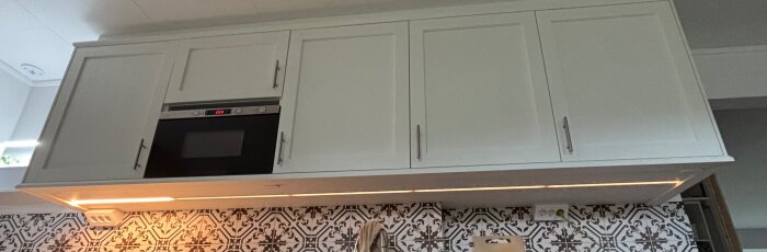 Vitt köksskåp med inbygd mikrovågsugn ovanför mönstrat kakel och köksbänk.