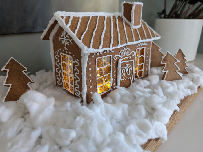 Pepparkakshus med vitt kristyrdekoration och snötäckta granar. Fönstren lyser av ljus inuti, och ett bomullslandskap omger det.