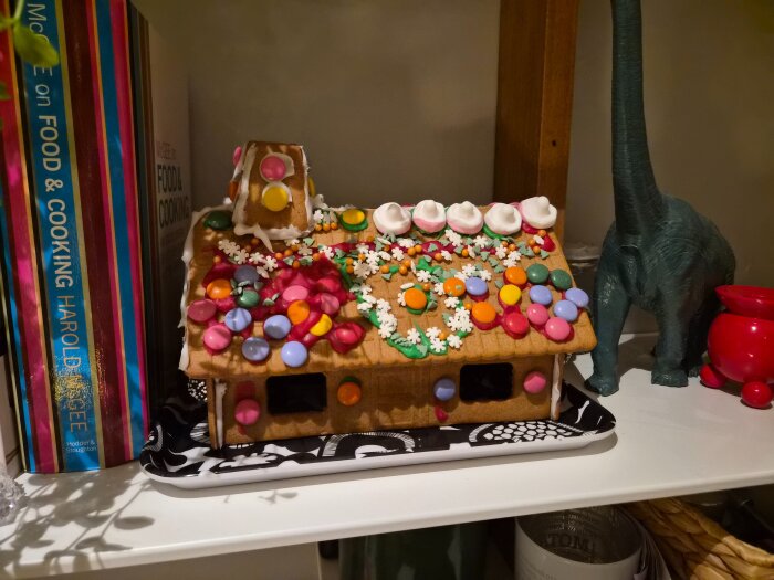 Pepparkakshus dekorerat med färgglada godisar och glasyr, omgiven av böcker och en dinosaurieleksak.