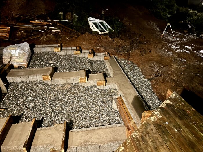 Grundläggningsarbete med betongblock och trästöd på en grusig yta, omgiven av jord och byggmaterial i bakgrunden.