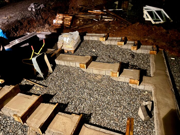 Byggarbetsplats med grusmark och formade betongblock, en skottkärra och staplade byggmaterial. Större objekt i bakgrunden.