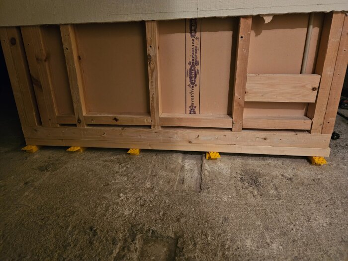 Väggens trästomme är uppallad med gula plastkilar på ett betonggolv.