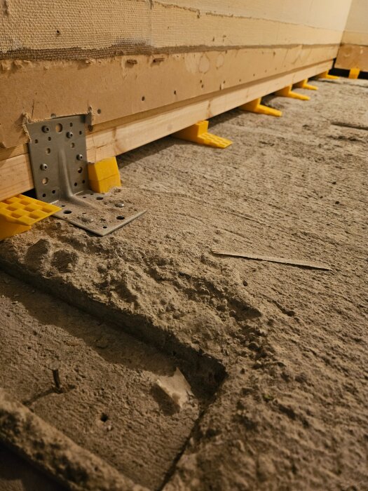 Vägg upplyft på gulnade plastkilar över betonggolv; L-fästen syns på träets kant.