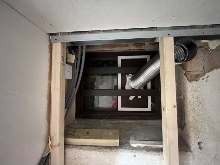 Ventil med silverfärgad rördragning i en halvt färdig vägg, omgiven av isolering och träreglar. En improviserad täckskiva ses också.