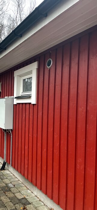 Rött trähus med vitt fönster och ventilationsgaller under ett takfot. Utomhusmiljö med träd i bakgrunden.