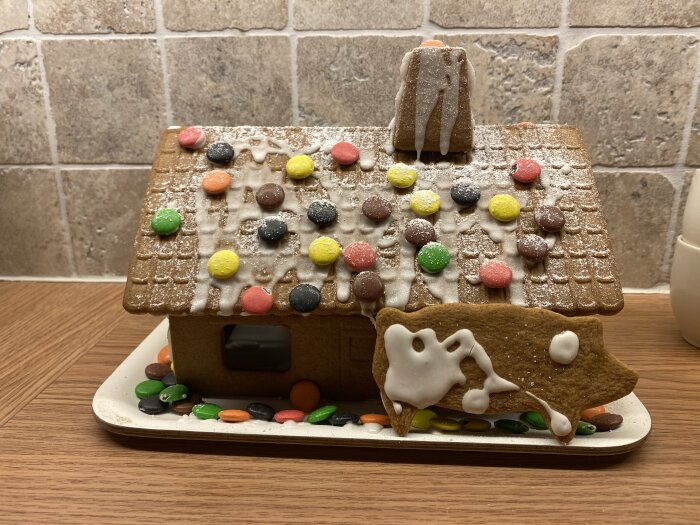 Pepparkakshus dekorerat med glasyr och färgglada godisar på taket, stående på en tallrik i köket.