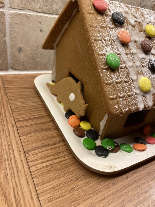 Pepparkakshus med glaserat tak och dekorerat med färgglada godisar, stående på en vit platta i ett kök.