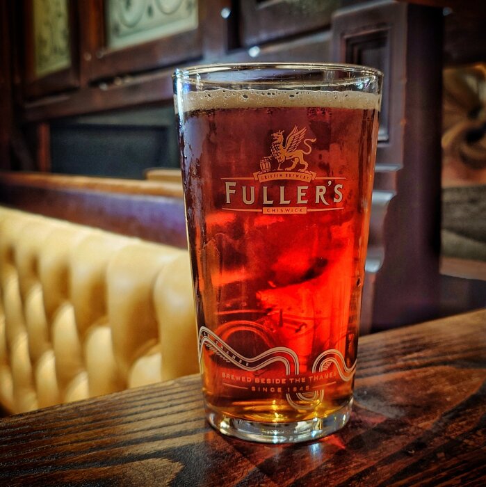 Ett glas med mörk öl från Fuller's på ett bord i en pubmiljö med gula lädersoffor i bakgrunden.