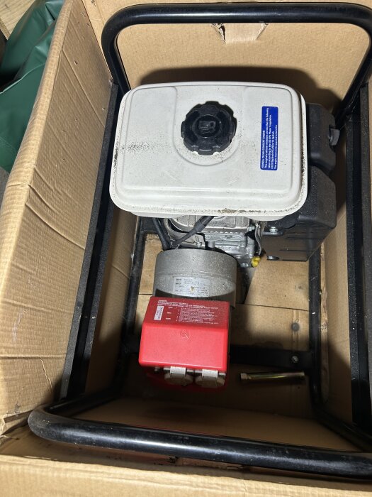 En generator packad i en kartong med en vit bränsletank och en röd elektrisk enhet.