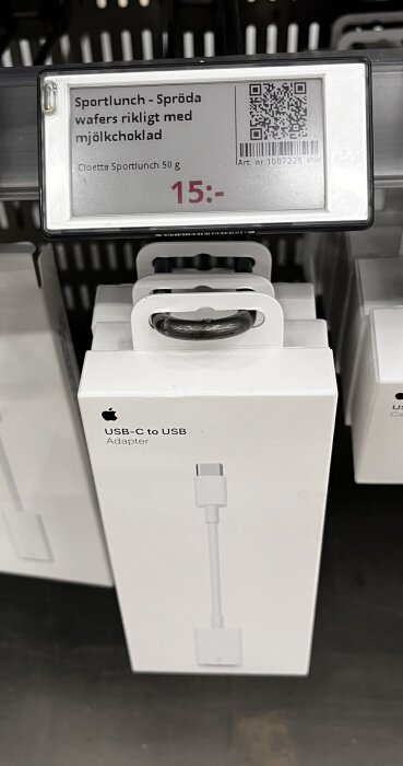 Förpackning med Apple USB-C till USB-adapter, placerad i butikshylla under prisetikett för chokladkaka, läser 15 kronor.