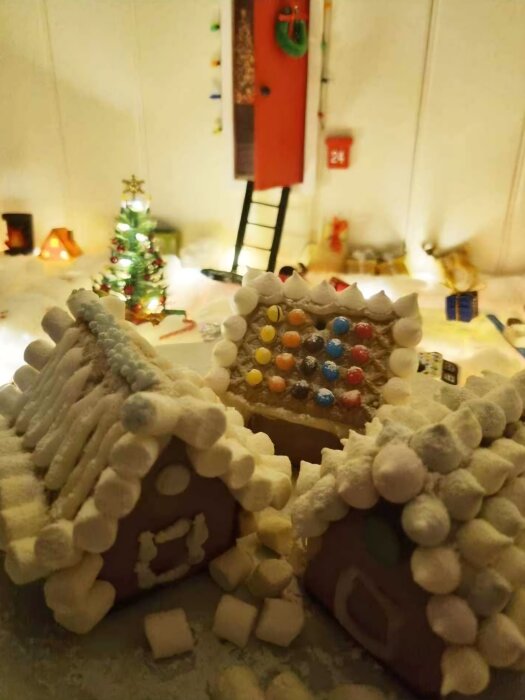 Tre pepparkakshus dekorerade med godis och marshmallows, med en julgran och tomtens dörr i bakgrunden.