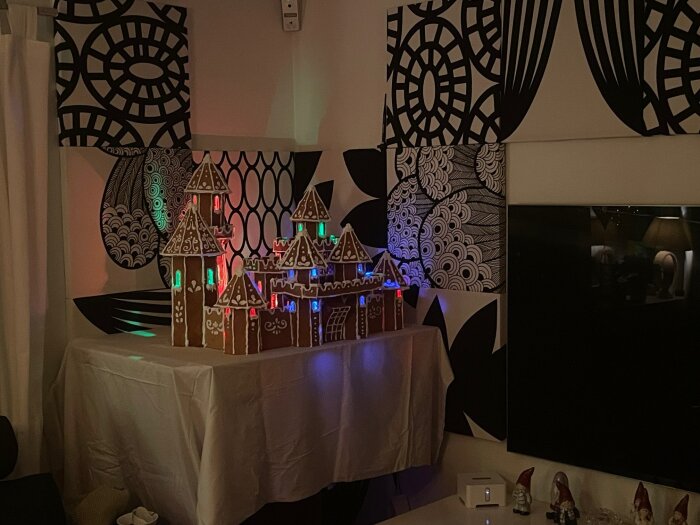 Dekorerat pepparkaksslott med färgglad ljusslinga på ett bord, omgiven av mönstrade tavlor och tomtar i ett rum.