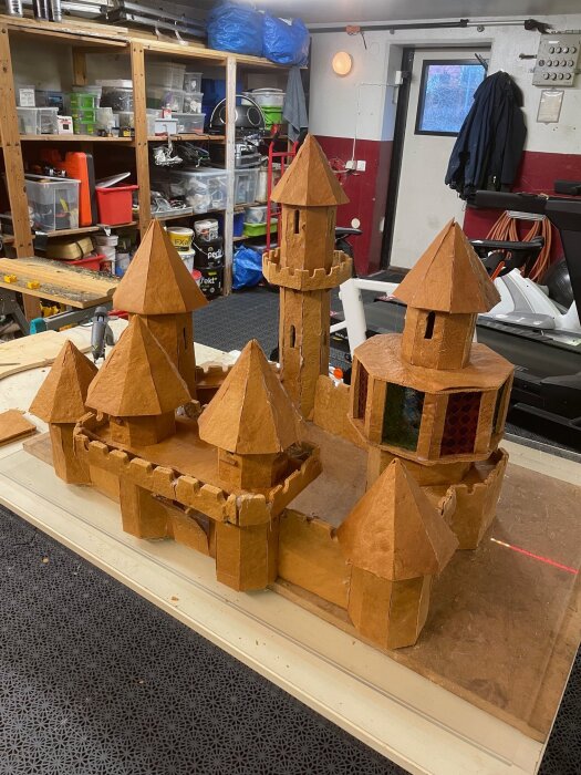 Pepparkaksslott på byggbord med tinnar och torn, gjort av 7,5 kg pepparkaksdeg, i ett verkstadsutrymme med hyllor och verktyg i bakgrunden.