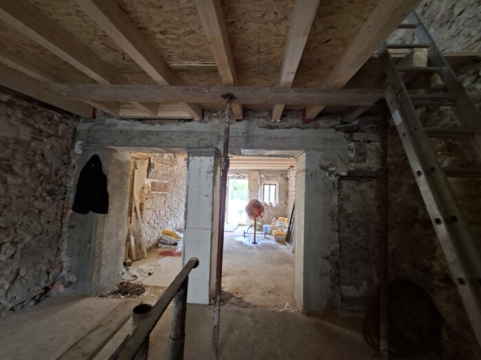 Renovering av hus med öppning mellan kök och vardagsrum, synliga takbjälkar och stege; byggmaterial och verktyg i bakgrunden.