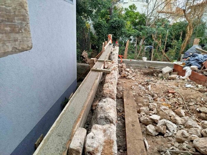 Stenmur under konstruktion med återanvända stenar från uppbrutna väggar, belägen intill en trädgård med gröna buskar i bakgrunden.