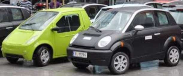 Två elbilar, en grön och en svart, står parkerade bredvid varandra på en rad med andra bilar.