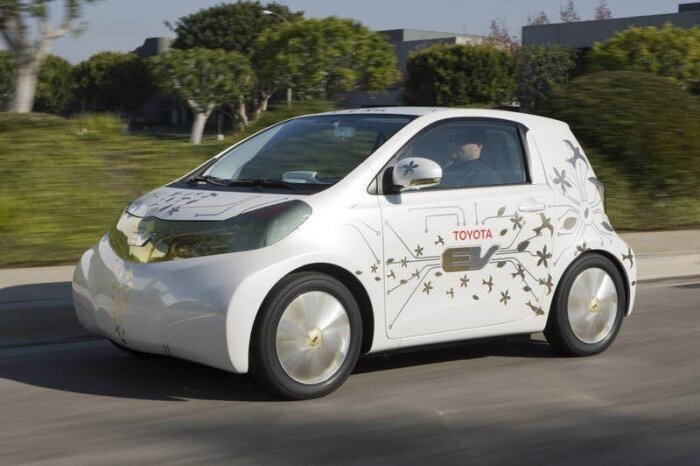 En liten vit elbil med blommönster och "Toyota EV" text på sidan, kör längs en väg omgiven av träd och buskar.