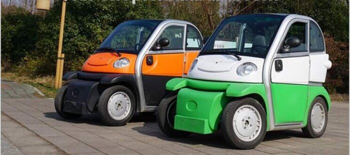 Två små, färgglada elbilar, en orange och en grön, står parkerade på en trottoar framför en grönskande bakgrund.