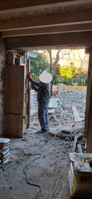 Person arbetar på tröskel mellan kök och vardagsrum, med byggmaterial och stenmur synliga genom öppningen mot trädgården.