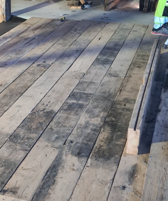 Trägolv i gammal lada med breda, slitna brädor och synliga mellanrum. Golvet är delvis återställt med bevarad patina från ursprungliga brädor.