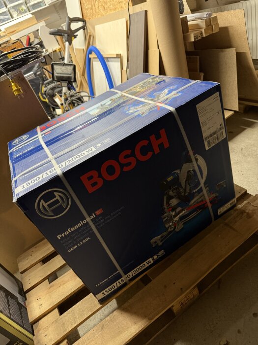 Kartong med Bosch kap- och gersåg på en lastpall i en verkstad fylld med byggmaterial.