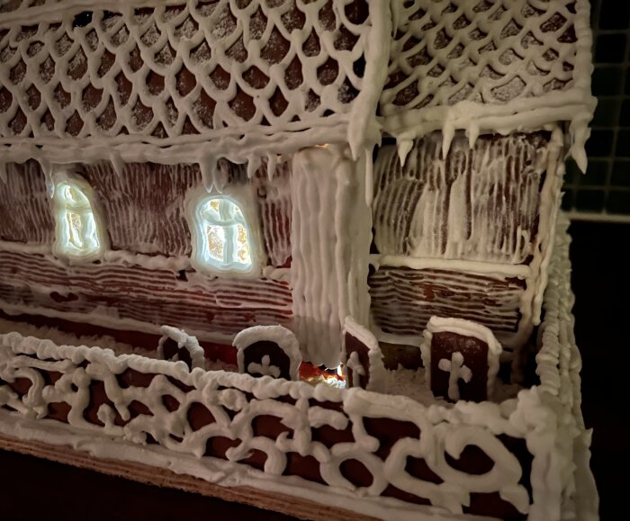 Pepparkakshus med detaljer av Dalarö kyrka, dekorerat med glasyr och upplysta fönster, samt ett staket med snörik mönster framför.