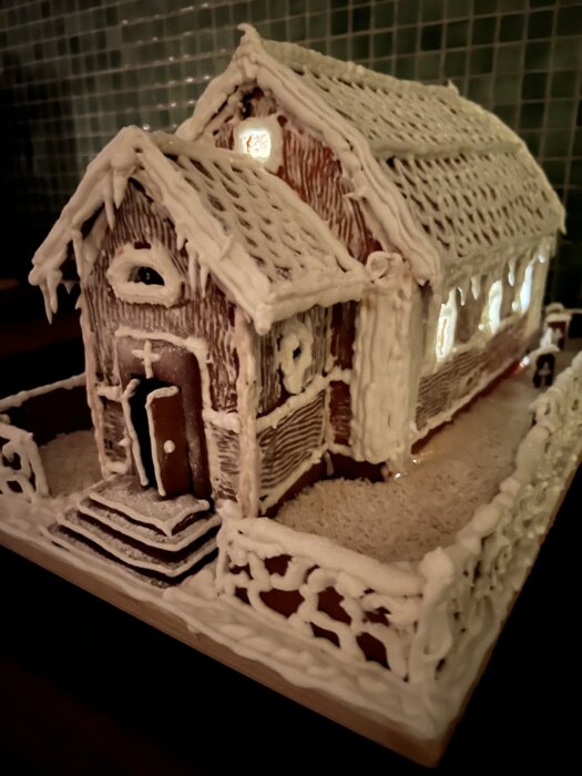 Pepparkakshus inspirerat av Dalarö kyrka, med detaljer av glasyr som imiterar snö och ljus inuti, står på en bricka med staket av pepparkaka.