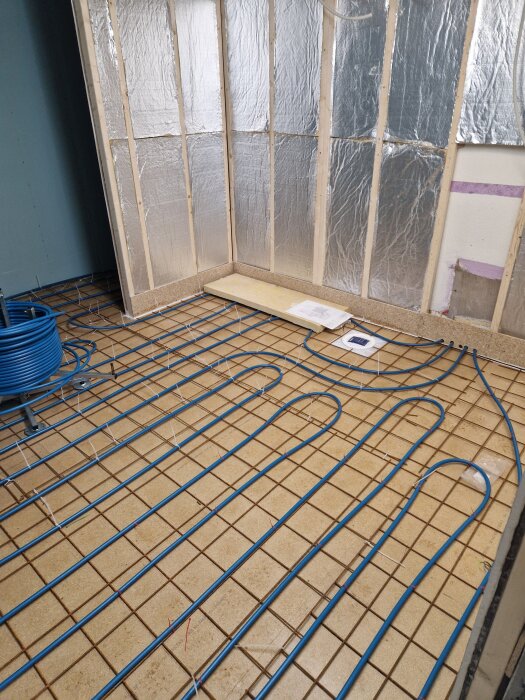 Golvvärmeslingor i blått rör på galler i byggprojekt, ofärdigt rum med isolerade väggar i bakgrunden.