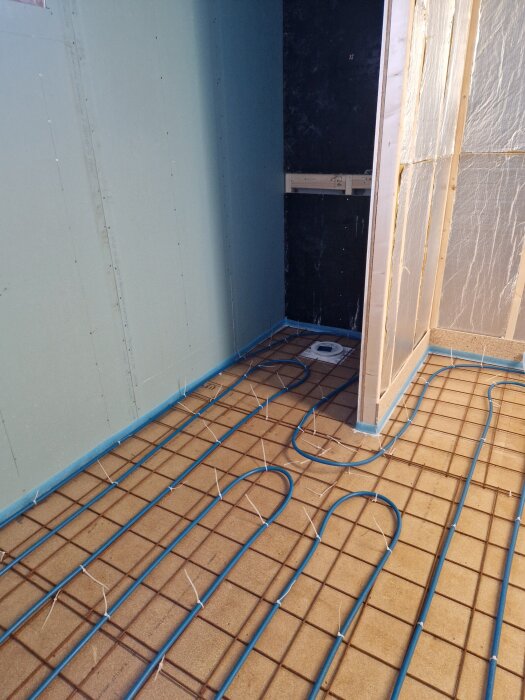 Golvvärmeslingor i blå plast på ett cementgolv i ett pågående badrumsbygge, med isolerade väggar omkring.