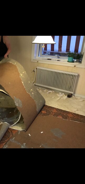 Person river upp bruna mönstrade golvmattor i ett äldre hus, under en lampa och framför ett fönster med persienner.