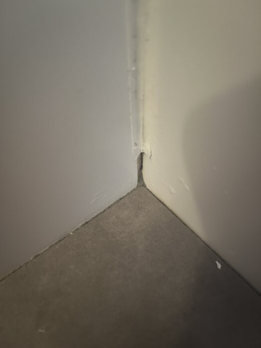 Hörn av ett rum med spricka i väggen och ojämnt fogmaterial mellan väggen och golvet i ett renoveringsprojekt.