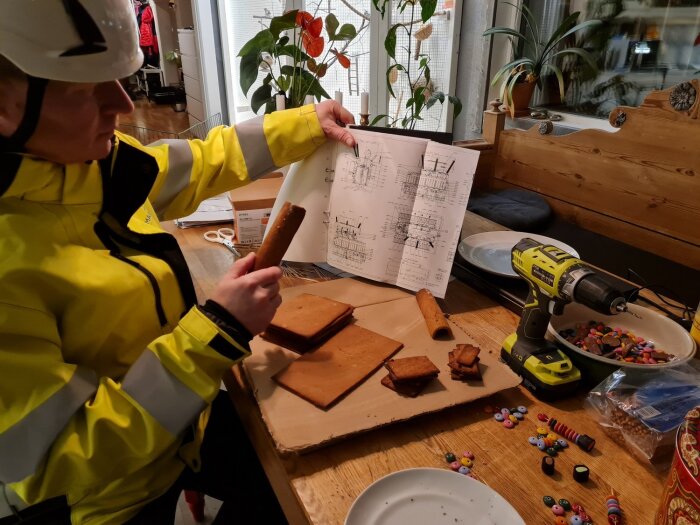 Person i säkerhetsutrustning bygger en modell med kakor och godis enligt ritning, med en borrmaskin på bordet bredvid.