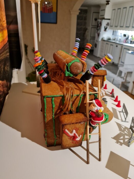 Pepparkakshuskonfiguration av en transformatorstation med godisledningar, tomtefigur på stege och detaljer av lakrits och choklad i ett köksmiljö.