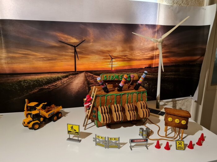 Modell av ett transformatorverk i pepparkaka, med små skyltar och koner, framför en fond av en väg med vindkraftverk i solnedgång.