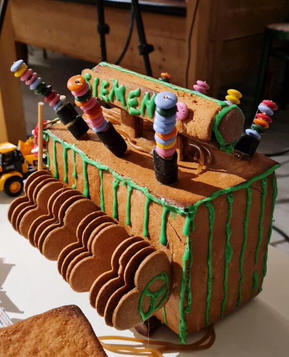 Pepparkakshus format som ett transformatorstation, dekorerat med godis och grön glasyr, med bokstäver på taket och kablar av lakritsremmar.