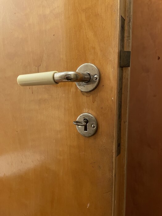 En trädörr med ett gammalt lås och en nyckel som sitter fast i låset, bredvid ett dörrhandtag med vit plasthandtag.