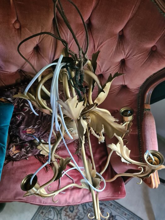 Guldfärgad ljuskrona i en rosa fåtölj med synliga kablar och lamphållare, diskussion om jordad kabel och ledare i inlägget.