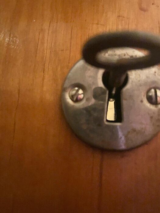 Nyckel fast i lås på gammal trätoalettdörr, närbild visar nyckeln i stillastående vridet läge och metallplatta runt nyckelhålet.