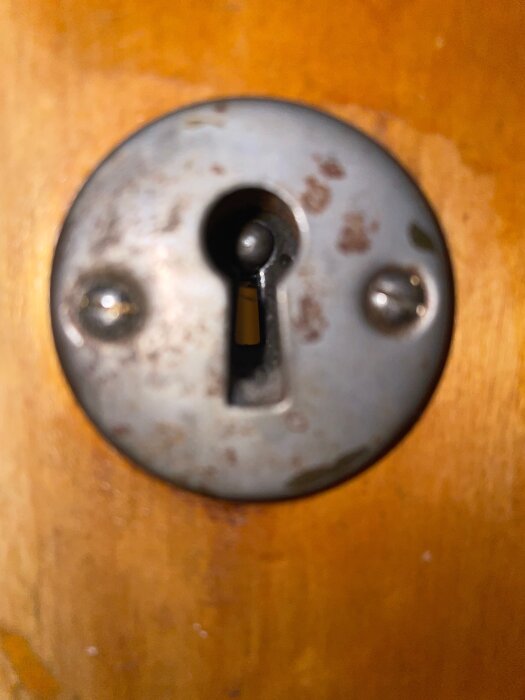 Närbild av en gammal nyckelskylt i metall med ett nyckelhål på en träfärgad dörr, delvis rostangripen.