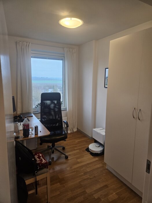 Arbetsrum med skrivbord, svart kontorsstol, och vy från fönster. Vit garderob till höger och liten tavla på väggen. Minimalistisk inredning.
