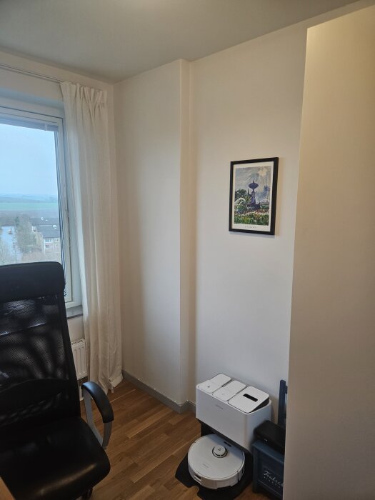 Arbetsrum med vit vägg och en tavla föreställande ett landskap, med en kontorsstol, fönster med vita gardiner och en robotdammsugare på golvet.